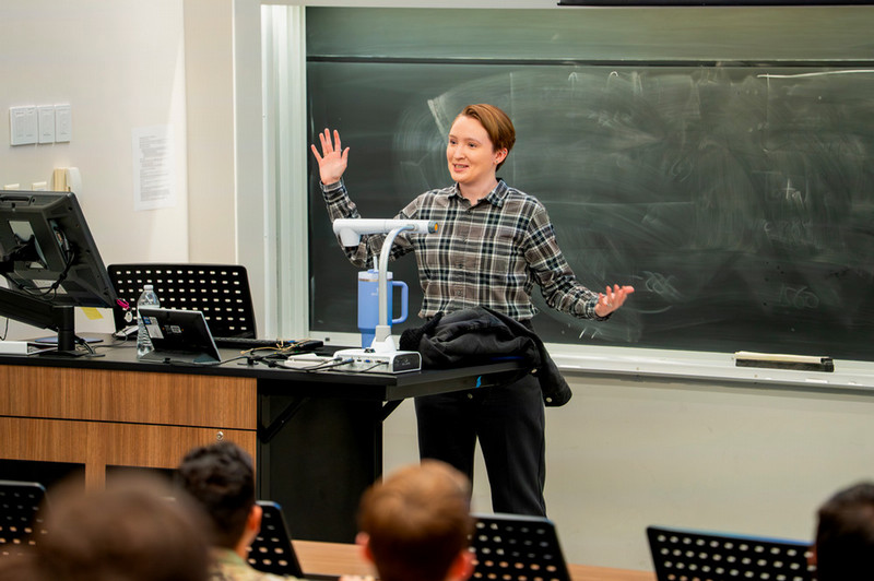 A picture of a woman in slacks and a plaid shirt presents in front of a crowd of students with her arms open wide. 